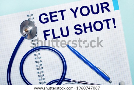 On a light blue background, an open notebook with the words GET YOUR FLU SHOT, a blue pen and a stethoscope. View from above. Medical concept
