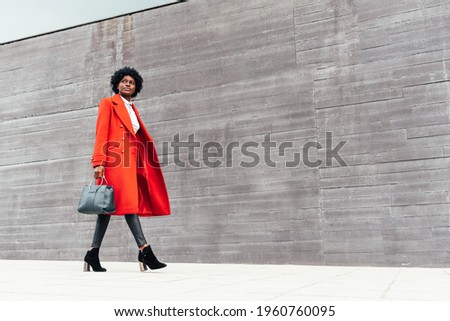 Fashionable black woman in red coat walking on out of focus background. Stylish African American woman in bright red jacket, black pants and handbag walking down the street. copy space photography
