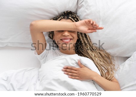 Top view of a pretty african-american young girl hiding her face with a hand in her bed. Cheerful black woman covering eyes with arms while wake up happy in the morning.