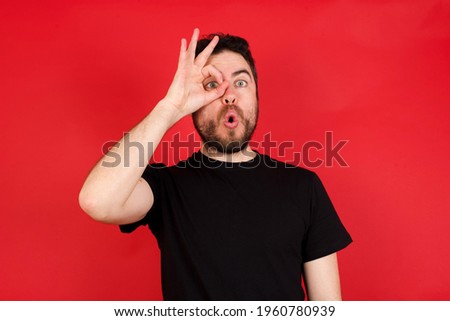 Young caucasian man wearing black t-shirt over red background doing ok gesture shocked with surprised face, eye looking through fingers. Unbelieving expression.