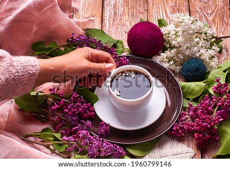 Hand holds a cup of morning coffee with spring lilac flowers branches blossoming on wooden background view from above. Flat lay underground style. Expensive colors. Creative design of flowers.