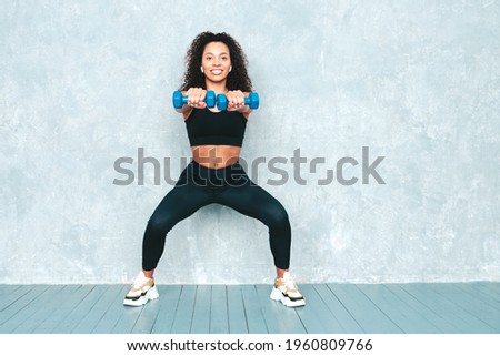 Fitness smiling black woman in sports clothing with afro curls hairstyle.She doing squats. Young beautiful model with perfect tanned body.Female holding dumbbells in studio near gray wall