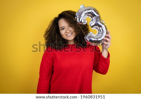 Young african american woman wearing red sweater over yellow background holding a number three balloon. Celebration concept