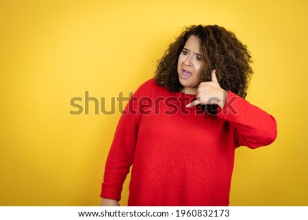 Young african american woman wearing red sweater over yellow background confused doing phone gesture with hand and fingers like talking on the telephone