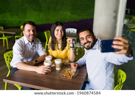 Attractive friends and co-workers taking a selfie with a smartphone while sitting together at a table in the food court and eating lunch after work
