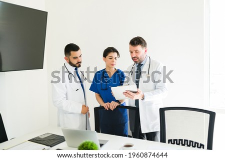 Latin hospital director showing the medical information of a patient on a tablet to a young doctor and female nurse at the meeting room Royalty-Free Stock Photo #1960874644