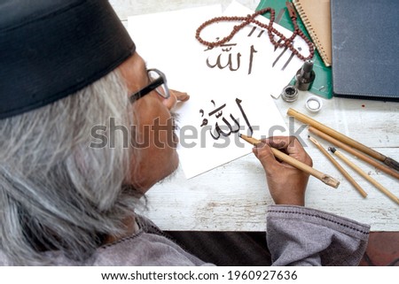 Senior Muslim man practicing  handwriting Khat with bamboo pen on paper, Arabic letters mean the name of Muslim god "Allah"