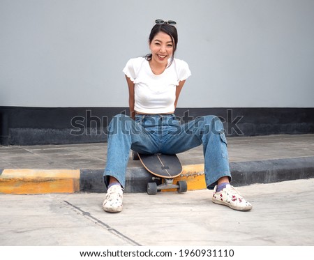 Asian girl, skateboarder sitting on sidewalk relaxing after exercise. Young beautiful woman sits on skateboard happy smiling. Surf skate is trendy outdoor extreme sport in Thailand. Copy space