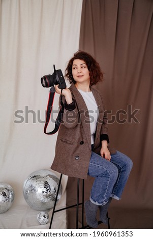 brunette woman with curly hair in a jeans and a checkered jacket sits on a high chair in a photo studio with a camera in hand. photographer's job or favorite hobby