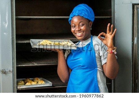 close up image of beautiful african lady in front of a local oven- cheerful black woman in apron and a head wrap with cookies - baking concept Royalty-Free Stock Photo #1960977862