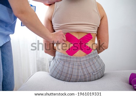 Physical therapist applying kinesio tape on female patient's lower back. Kinesiology, physical therapy, rehabilitation concept. Female loin with physio tape on light background