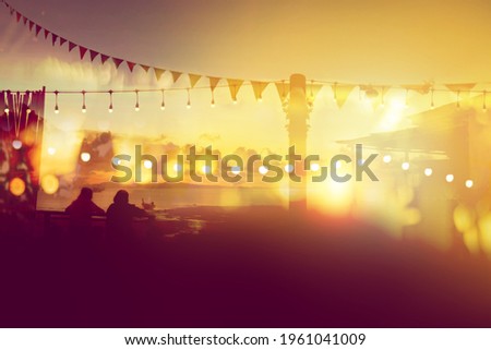 two friends watching sunset at restaurant on the beach, blurred bokeh light on sunset with yellow string lights decor in beach restaurant 