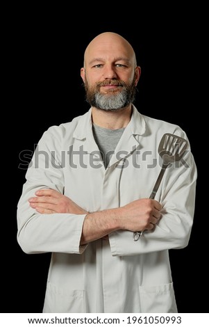 Portrait of handsome chef cook on a black background. The model is in his 40s, bald with grey beard wears white uniform. Slim body type. Cooking industry professional. Holding metal turner