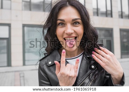Attractive Caucasian young brunette brunette woman in leather jacket shows tongue and gesture of rocker, punk or horns on street in city on autumn day. Metal music concept.