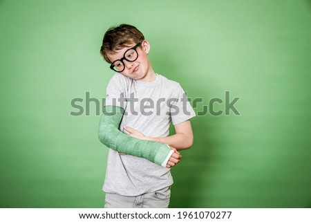 cool young schoolboy with black glasses and broken arm with green plaster standing in front of green background