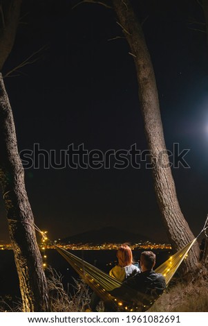 Couple relaxing in a hammock and enjoying starry sky. View from the top of the mountain to the lights of the city of Naples. Volcano Vesuvio. Sea and romance. Vertical photo. Sorrento Italy