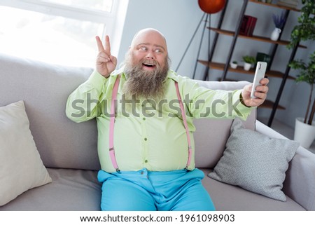 Portrait of positive cheerful guy sit on sofa take selfie fingers show v-sign beaming smile free time indoors