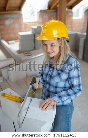 pretty young female bricklayer with yellow safety helmet is sawing bricks on a construction site in the house and is happy