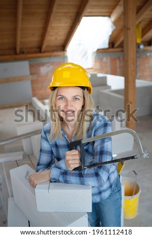 pretty young female bricklayer with yellow safety helmet is sawing bricks on a construction site in the house and is happy