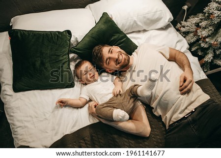 Portrait of a young beautiful family in beige T-shirts lying on a white bed in a dark interior. Husband and son. Dad and baby. Entertainment at home, spending time together. Love, care. Men and child.