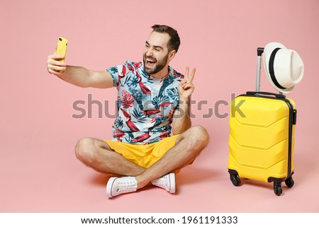 Full length funny young traveler tourist man sit on floor doing selfie shot on mobile phone showing victory sign isolated on pink background. Passenger travel on weekend. Air flight journey concept