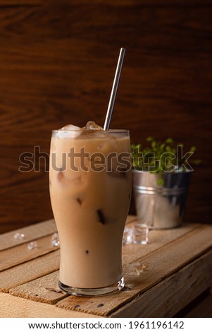 In a glass filled to the top with ice and coffee with milk. A glass of iced coffee sits on a crate against a dark wooden background.