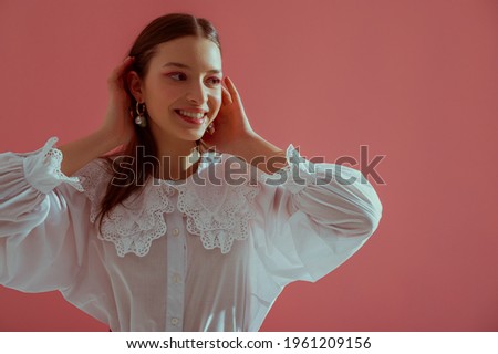 Fashionable  happy smiling brunette girl wearing trendy white vintage cotton blouse with lace collar, stylish pearl earrings posing on pink background. Copy, empty space for text