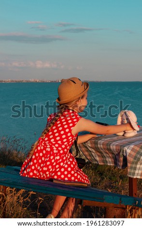 Little photographer child in straw hat and red polka-dot dress on vintage bench taking picture of soft pink rabbit toy on sea lanscape background. Girl looks at camera in hands. Friendship fun travel.