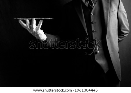 Portrait of Butler or Waiter in Dark Suit and White Gloves Expertly Holding Silver Tray on Black Background. Concept of Service Industry and Professional Hospitality.  Royalty-Free Stock Photo #1961304445