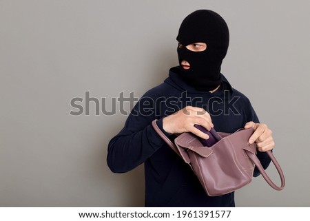 Insidious male villain in balaclava stands with stolen women's bag and wallet, afraid that he will be caught, turns around and looks carefully back, isolated over gray background.