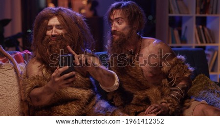 Wild bearded man in animal savage fur taking selfie picture on smartphone camera interacting technoogy watching TV. His friend returning back home. Humor concept.