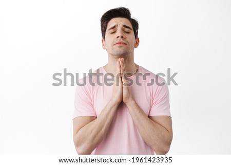 Hopeful worried man praying, close eyes and plead, anticipating results, standing nervous against white background in casual t-shirt