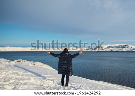 Portrait from back of warmly dressed woman in defocus standing in front of sea looking far away looking at winter rough water in slow motion.