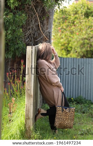 Fashion image of plus size woman wearing brown cardigan and leopard skin bag leaning against wooden fence. No face visible.