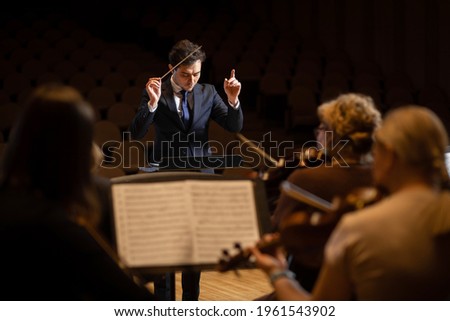 Conductor of symphony orchestra with performers in background in concert hall Royalty-Free Stock Photo #1961543902