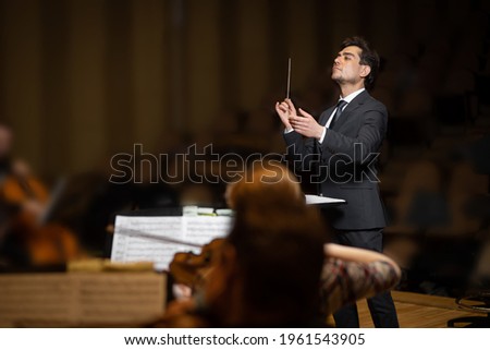 Conductor of symphony orchestra with performers in background in concert hall Royalty-Free Stock Photo #1961543905