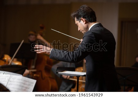 Conductor of symphony orchestra with performers in background in concert hall Royalty-Free Stock Photo #1961543929