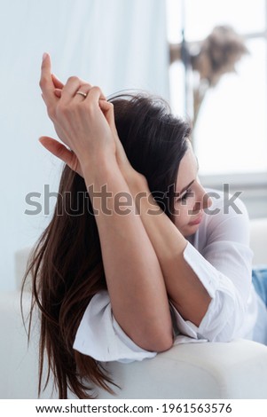 The brunette girl bowed her head on her hand and depicts sadness, perplexity, sits on a light sofa on a white background Royalty-Free Stock Photo #1961563576