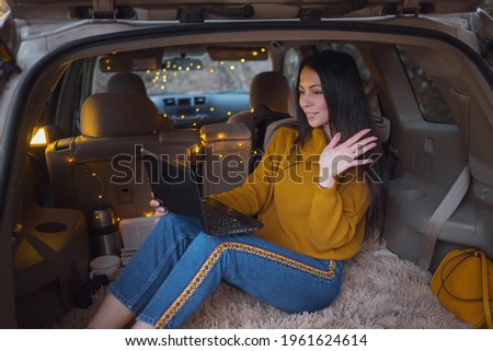 Young beautiful brunette enjoys relaxing in the trunk of her car. A yellow garland and a beige plaid create coziness, Woman reading email, chats in messengers and talking on video conference on laptop