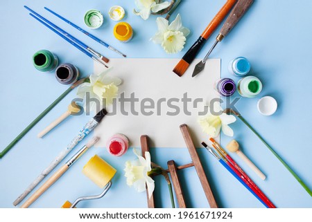various painting tools: brushes, spatula, roller, paints, wooden easel, spring flowers. the blank in the middle. flatley style on a blue background. copy space. art concept learning to draw in spring