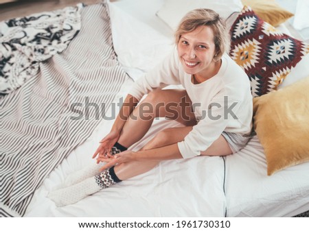 Young blonde female dressed in pajama sitting in the cozy bed with pillows and sincerely smiling and gazing at the camera. Staying home and sweet cozy home concept image