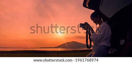 Silhouette of a woman holding a camera in the back of the car at sunset on the lake with copy space. Concept of Inspirational and positive thinking.