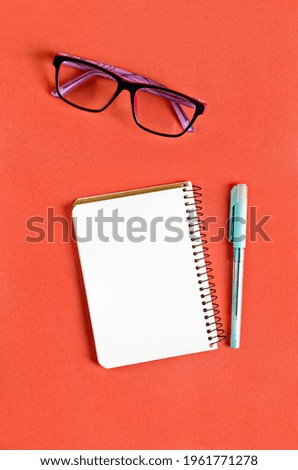 Blank white notebook, eyeglasses and pen on orange and green background. Minimalist concept with place for text. Mock up with decor elements.
