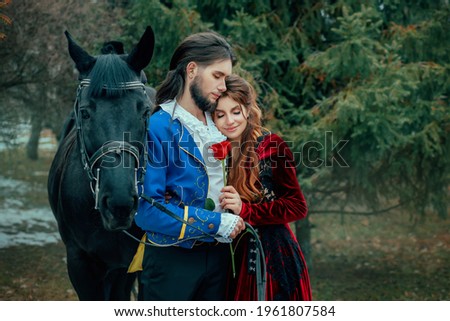 Medieval couple in love man and woman hugging in winter forest. Vintage clothing red long dress. Blue costume tailcoat caftan. Prince and princess together. Black horse. Art image redhead hairstyle