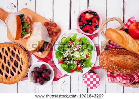 Summer picnic food table scene. Variety of cold salads, sandwiches, fruit and treats. Top view over a white wood background.
