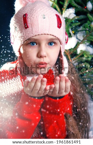 A girl child in a hat with a pumpon blows in the palm of her hand and snow flies out of them. A girl with real blue eyes. New Year's card. Background