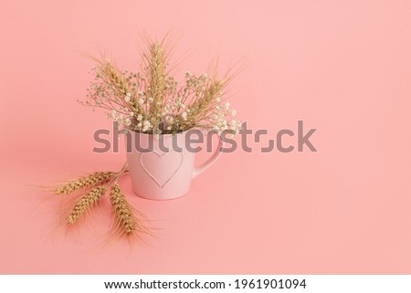 Pink mug with a picture of a heart and a bouquet of white flowers and ears of wheat. Wheat ears next to a mug on a pink background represent the israeli holiday of Shavuot