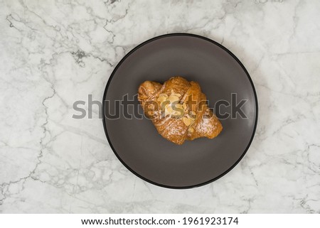 Flat lay view of gray plate with croissant on white marble tabletop.  Dark food photography of croissant on grey plate. The background is blurred.