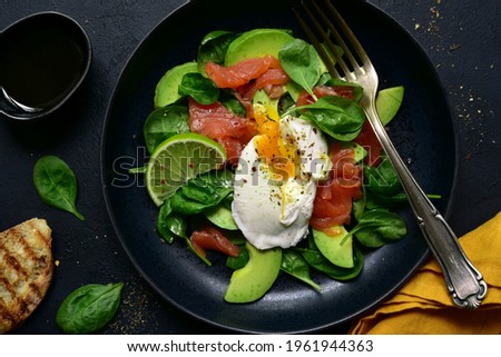 Avocado and baby spinach salad with salted salmon and poached egg on a black plate over dark slate, stone or concrete background. Top view with copy space.