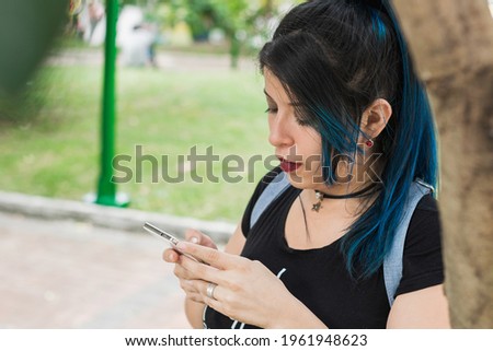 Beautiful young blue haired college girl looking at her social networks in a park leaning on a tree. girl looking at her cell phone in a park. surrounded by nature